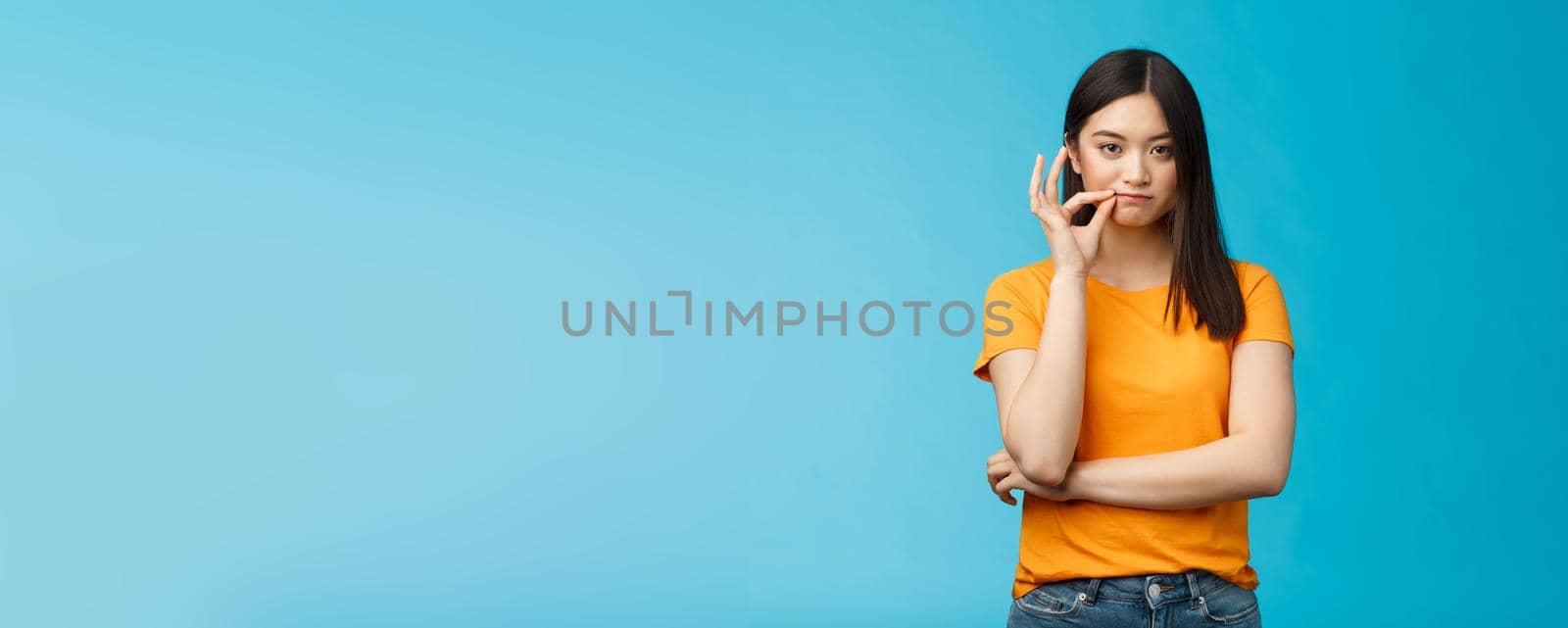 Serious-looking asian female friend promise keep secret, seal lips, hold zip near mouth look focused determined, stay silent and speechless, stand wearing yellow t-shirt blue background.
