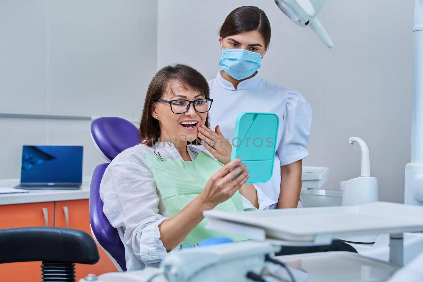 Happy middle aged woman together with dentist, patient sitting in dental chair looking at mirror. Prosthetics, treatment, implantation, dental teeth health, beauty care concept