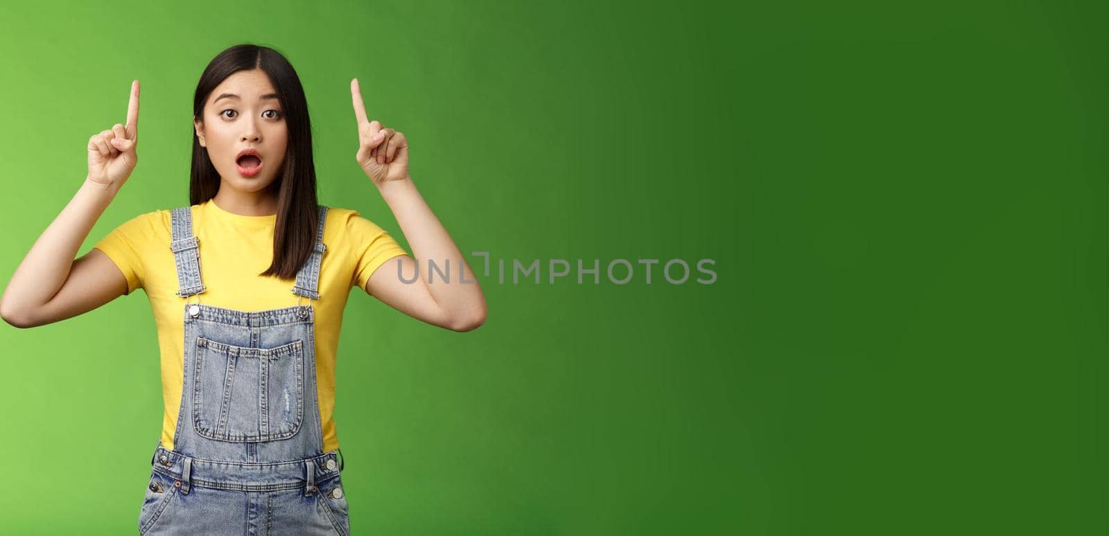 Impressed speechless shocked asian girl, pointing fingers up, drop jaw gasping astonished, stare camera amazed, telling about incredible prices new products, stand green background. Copy space