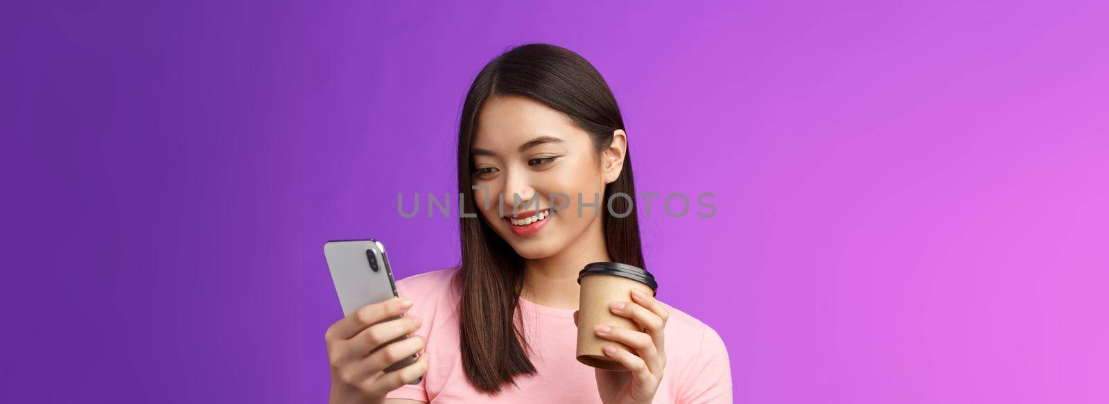Close-up tender friendly asian female blogger checking social media drinking take-away coffee, smiling joyfully look telephone screen, reading interesting article, texting, purple background.