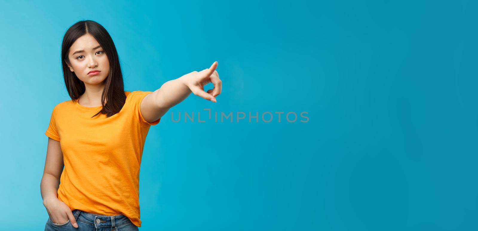 Go away from here. Serious-looking arrogant displeased bossy asian girl showing way-out, pointing sideways index finger look camera demand leave, stand blue background ignorant. Copy space