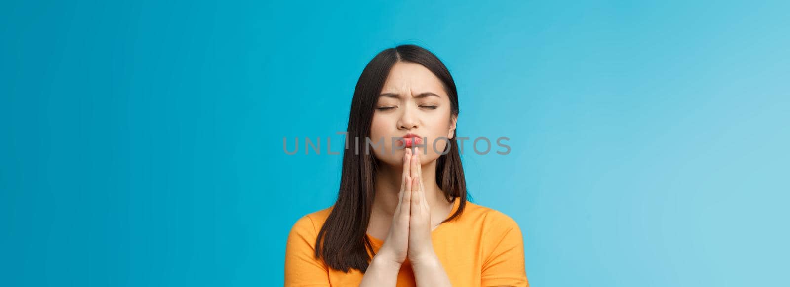 Determined motivated cute asian female praying dream come true, slap hands together pray pose, close eyes pouting eager win, achieve positive reply from university, stand blue background hopeful.