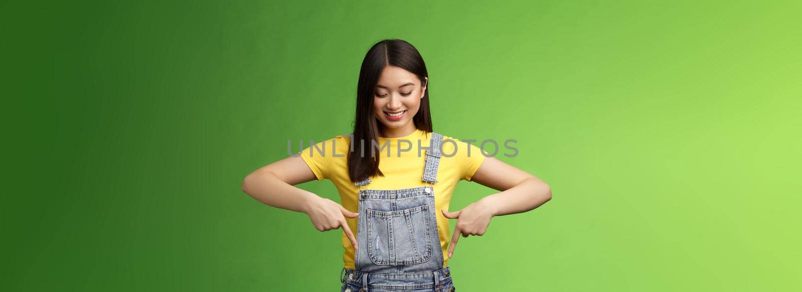 Studio shot easygoing cute asian female introduce new promo, pointing looking down amused pleasantly smiling, gladly show interesting thing, stand green background happy excited by Benzoix