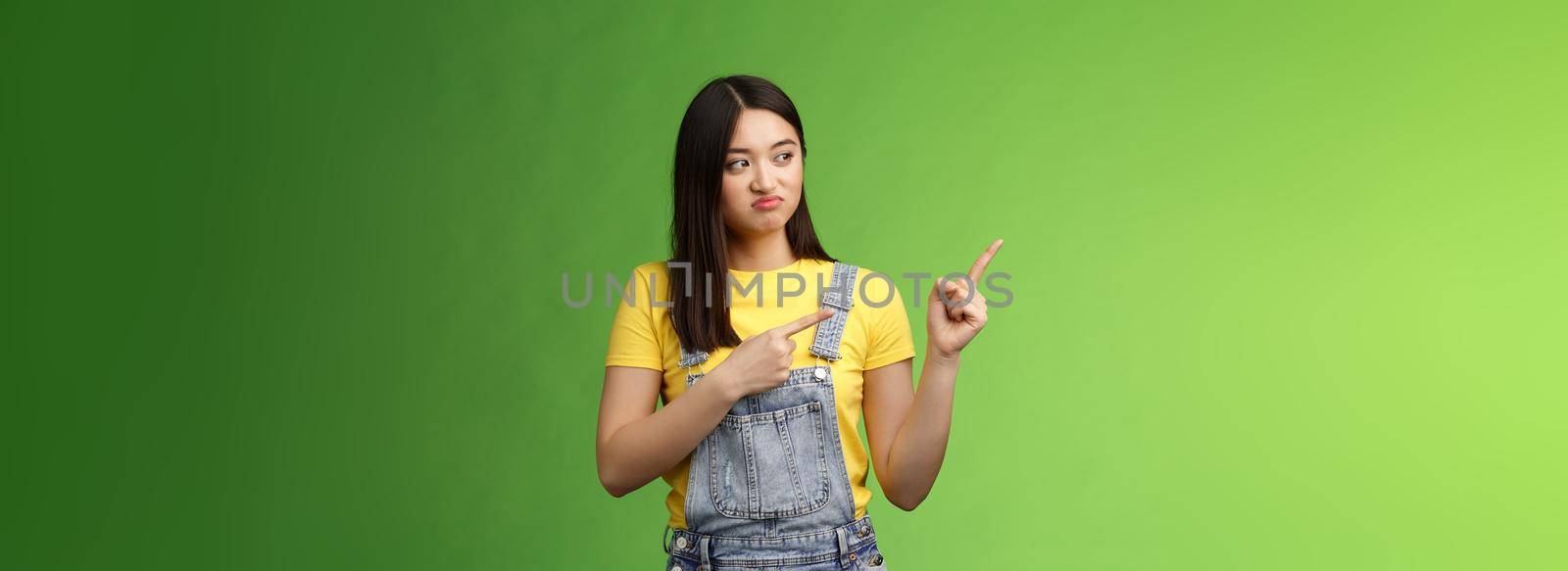 Upset cute asian female student pulling face regret shame, pity missing good chance buy product sale, pointing looking left disappointed, jealously gaze coworker, stand green background by Benzoix