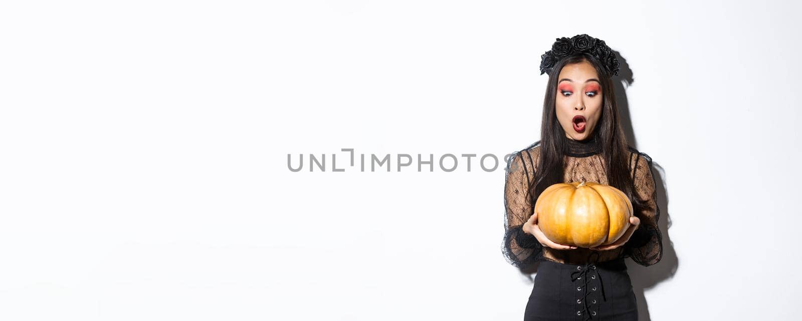 Image of amazed asian girl looking at big pumpkin fascinated, wearing gothic dress and black wreath, standing over white background by Benzoix
