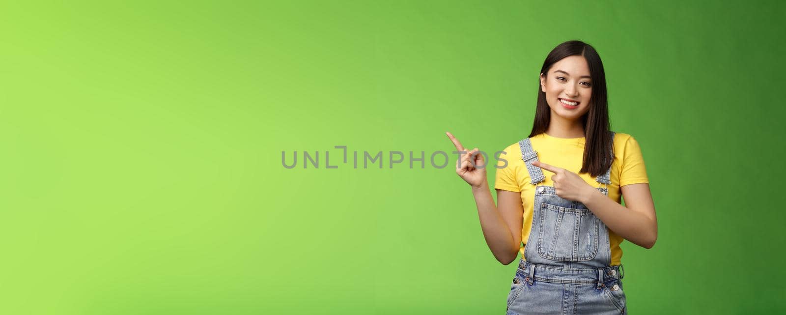 Lovely pretty tender asian woman showing way, tilt head flirty smiling, helpful pointing left, introduce plan, gladly present promo recommend cool new product, stand green background happy by Benzoix