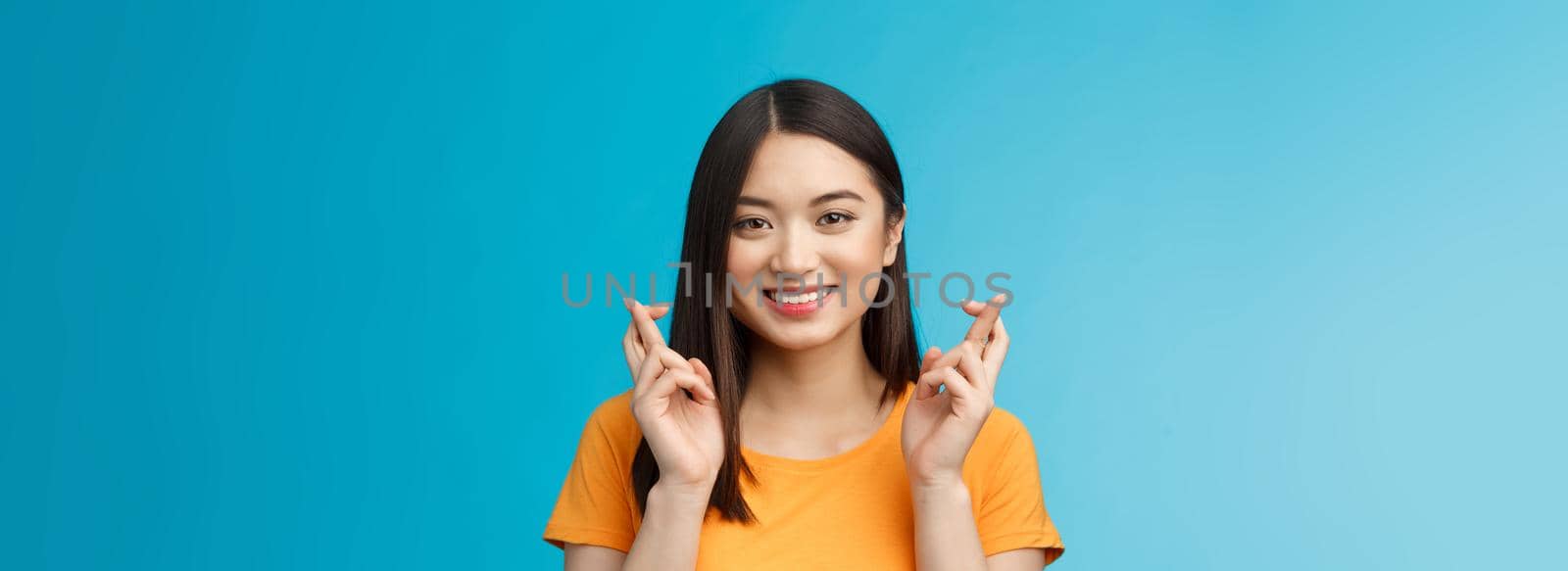 Close-up hopeful cute asian girl with short dark hair cross fingers good luck, praying smiling broadly awaiting positive news, faithfully hope win, stand blue background upbeat excited.