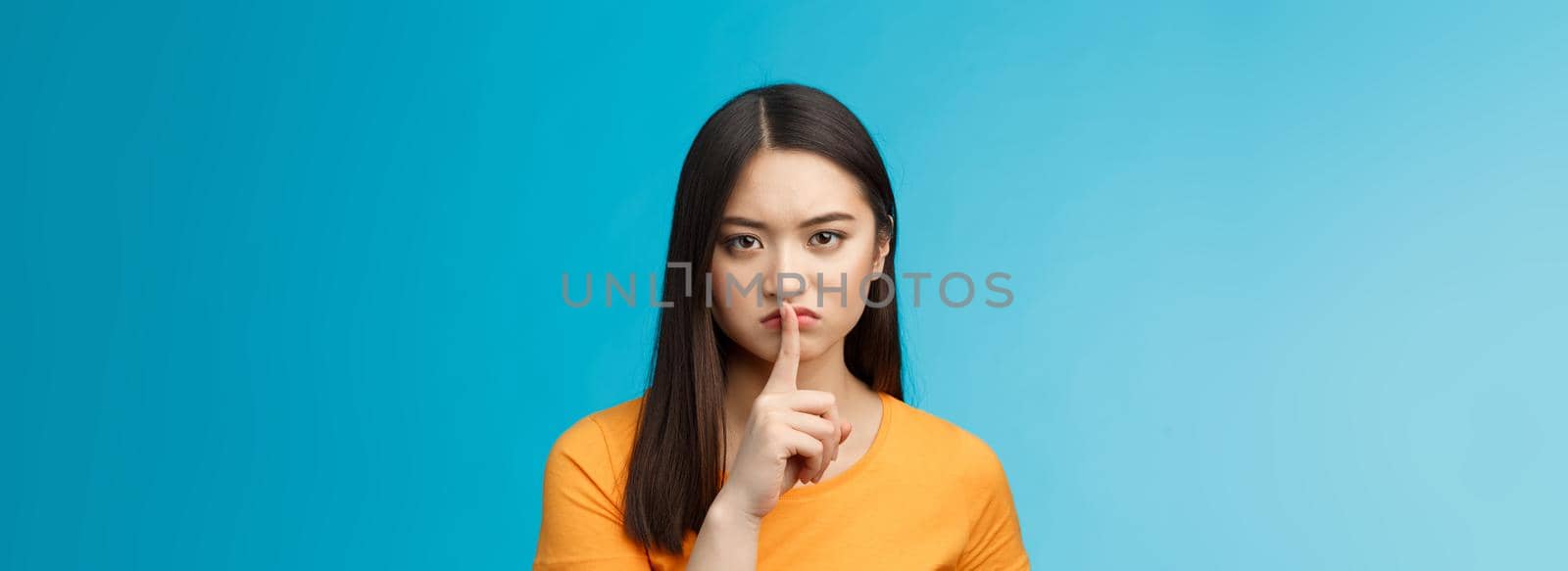 Hush stay silent. Serious-looking asian girl look displeased grumpy show shush sign press index finger lips, frowning upset, telling not speak, untell secret, prohibit loud music, blue background.