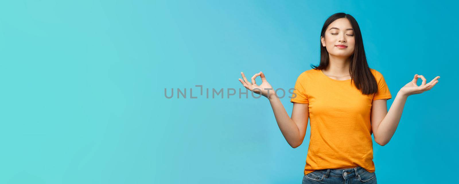 Peaceful charming relaxed asian girl buddhist meditating, breathing, inhale fresh air practice yoga, close eyes smiling relieved, stand lotus nirvana pose, reach zen, standing blue background.