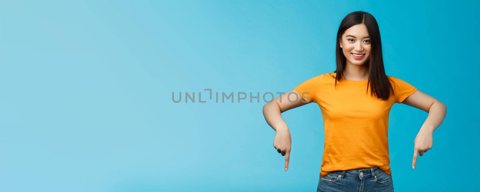 Cheerful self-assured asian girl dark haitcut wear yellow summer t-shirt introduce promo, pointing hands down, indicate bottom advertisement smiling delighted, share good link blue background.