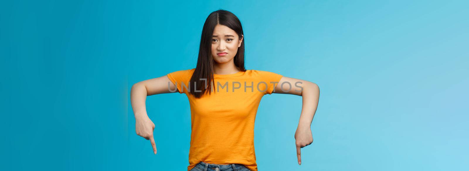 Moody sad asian brunette girl in yellow t-shirt smirking unsatisfied frowning bothered, complaining pointing down index fingers, showing bottom advertisement upset, jealous feel regret.
