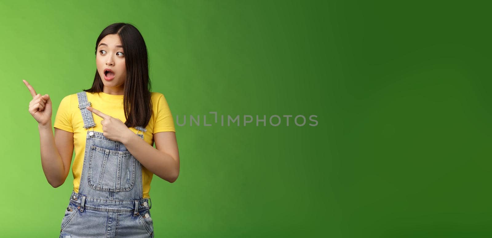 Shocked gasping alarmed pretty asian girl drop jaw impressed, turn pointing left hear loud bang, stand astonished speechless, gazing curious event, pose green background by Benzoix