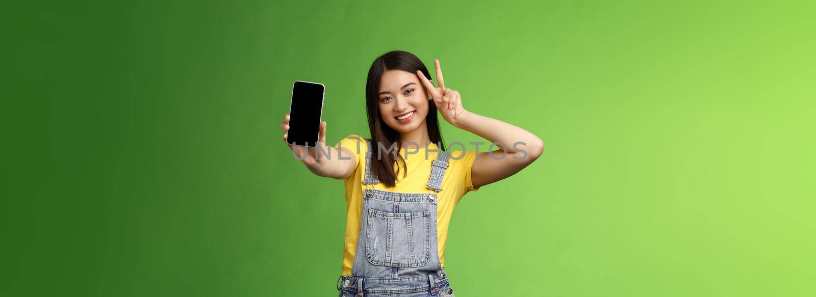 Cute carefree asian brunette showing application on smartphone screen, make victory peace sign, smiling joyfully, brag social media popularity, followers amount, stand green background by Benzoix