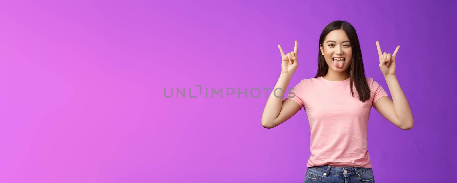 Rebellious amused asian girl enjoy rock-n-roll, attend awesome concert have fun, show tongue make heavy-metal gesture, express excitement and joy, stand purple background cheerful.
