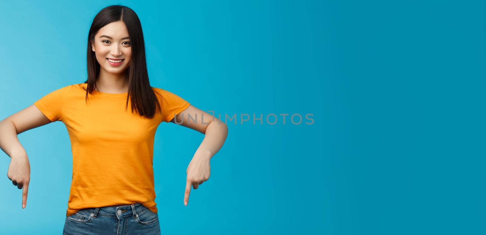 Cheerful self-assured asian girl dark haitcut wear yellow summer t-shirt introduce promo, pointing hands down, indicate bottom advertisement smiling delighted, share good link blue background by Benzoix