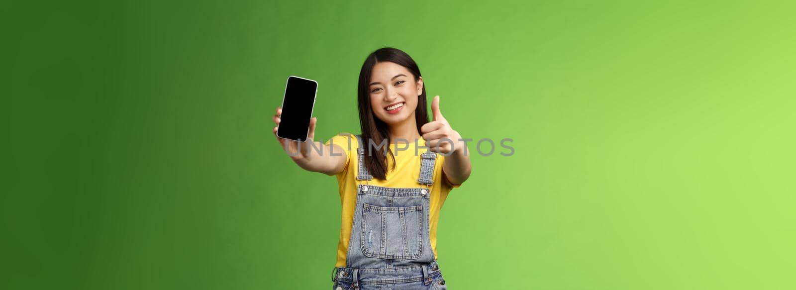 Cheerful satisfied cute asian brunette girl pleased awesome smartphone game, show telephone display, give thumb-up like sign, smiling broadly, introduce awesome online application, green background.