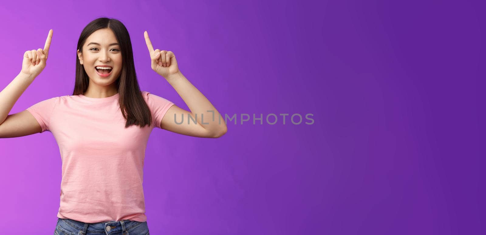 Charismatic good-looking lively smiling asian girl introduce product, pointing fingers up, indicate top advertisement, grinning toothy, excited telling good news, sharing link, purple background.