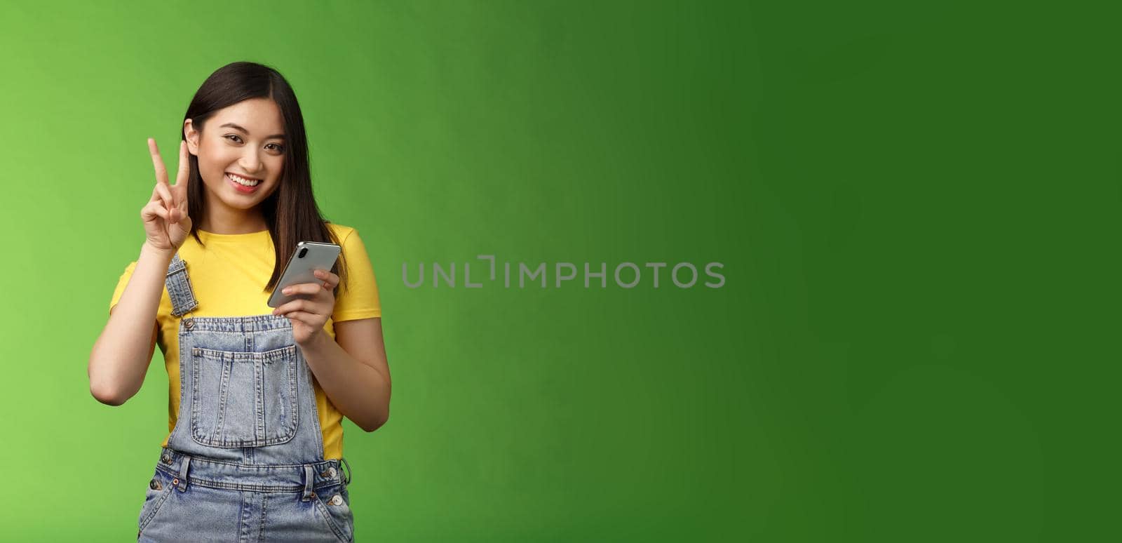 Cute tender asian woman hold smartphone, show victory peace sign, look camera delighted, carefree pose near green background, win online giveaway, triumphing joyfully beat score game by Benzoix