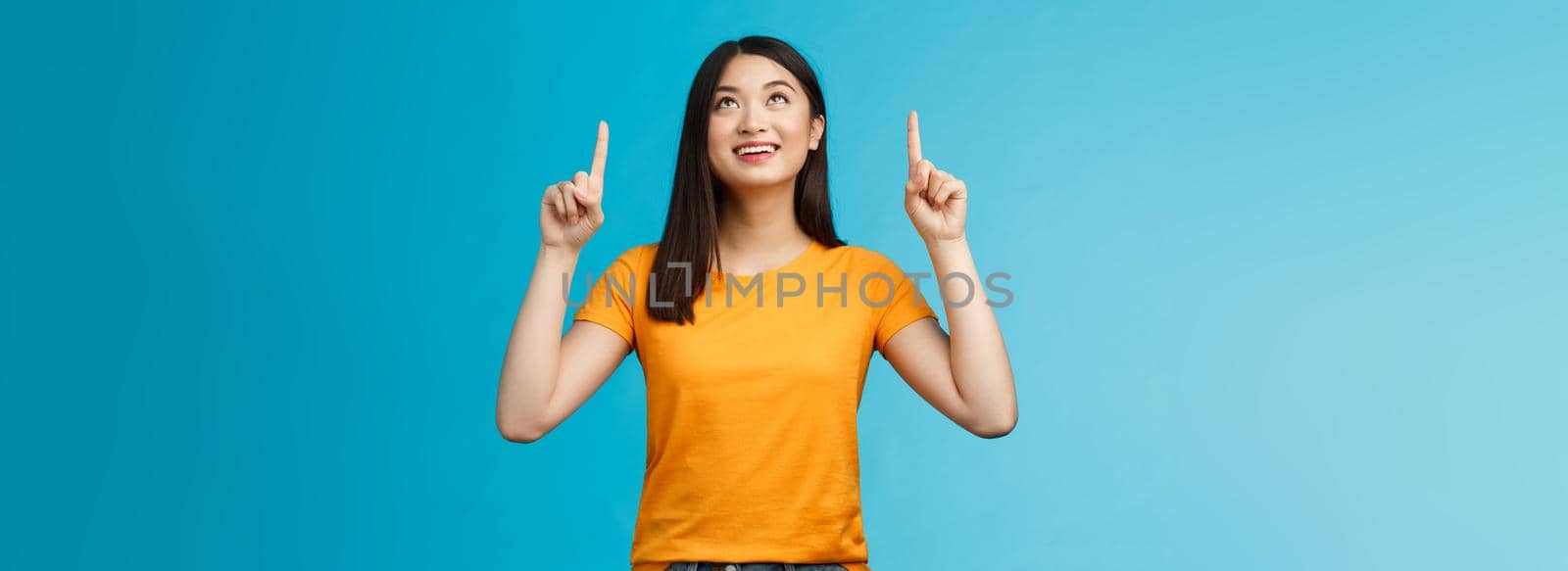 Asian girl contemplating view enjoy summer vacation like sunny days, look pointing up amused, feel enthusiastic upbeat, smiling broadly showing top promo, gaze with interest, blue background.