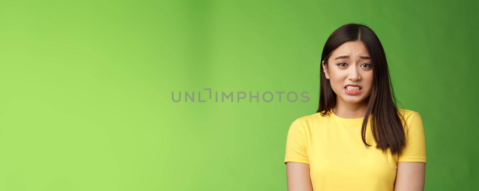 Anxious worried cute asian girl brunette clench teeth frowning shaking from fear look guilty afraid, stand nervous awaiting punishment, green background, apologizing ashamed by Benzoix