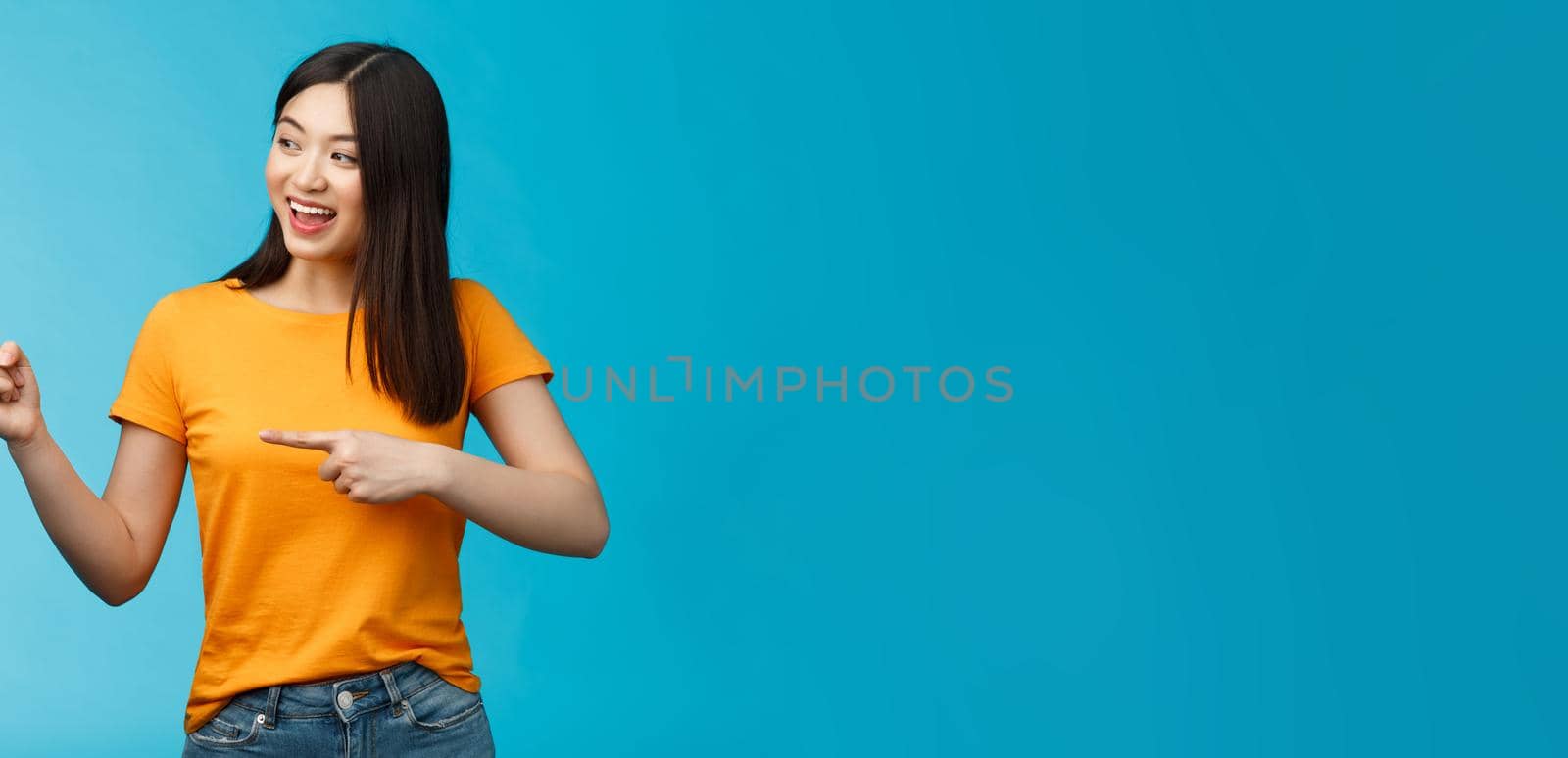 Friendly enthusiastic asian girl curiously looking sideways, pointing left interesting object, grinning joyfully discuss new opened store, stand blue background carefree lively talking.