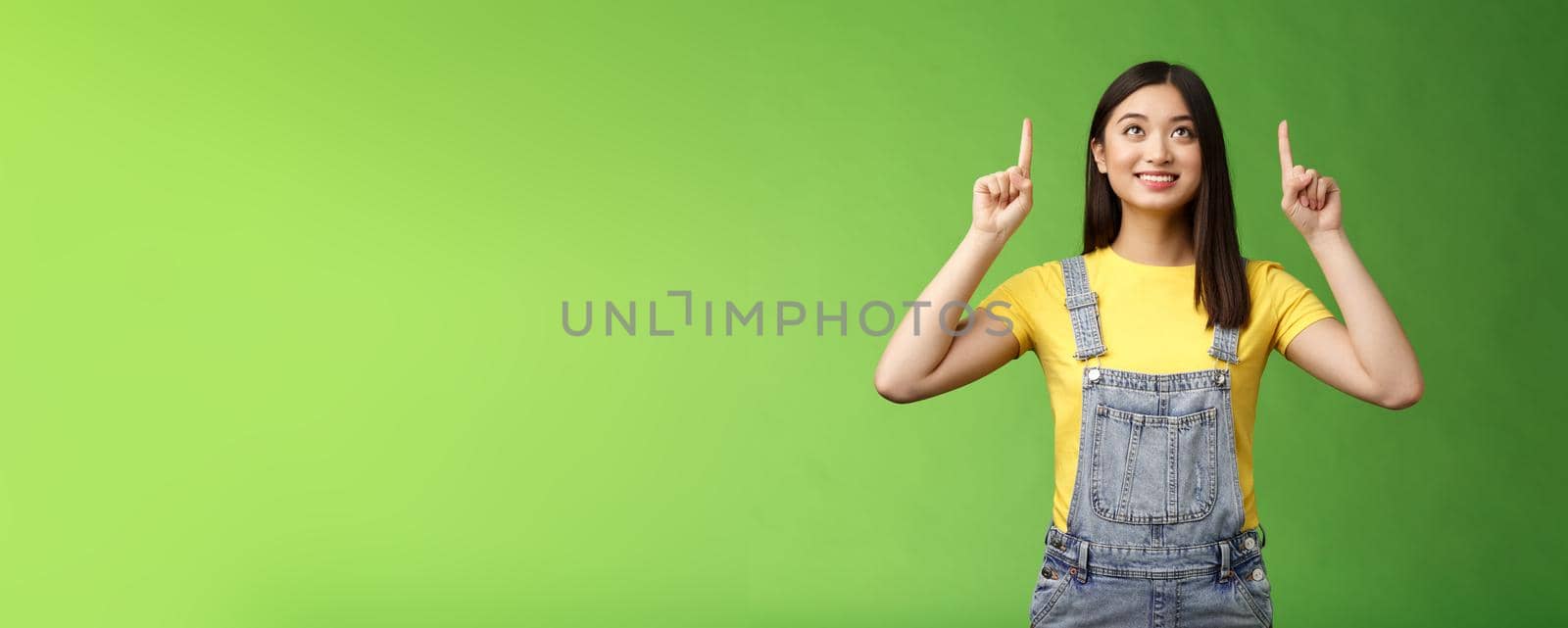 Dreamy hopeful cute asian girl look pointing up amused, smiling delighted, contemplate interesting object, enjoy stargazing, grinning satisfied, gazing top promo, stand green background.