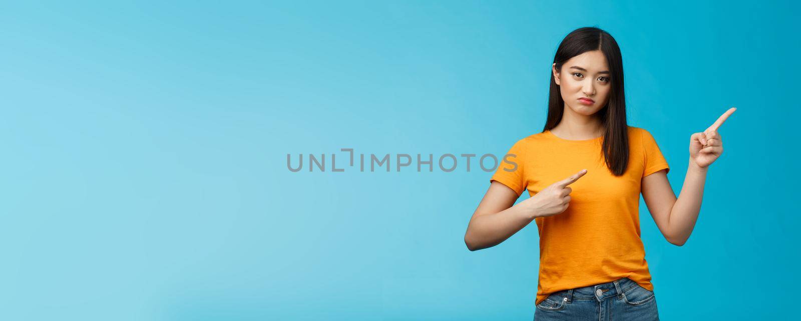 Picky disappointed asian girl with unsatisfied face, pointing upper right corner, sulking offended, feel jealous friend went summer vacation abroad alone, stand blue background displeased.