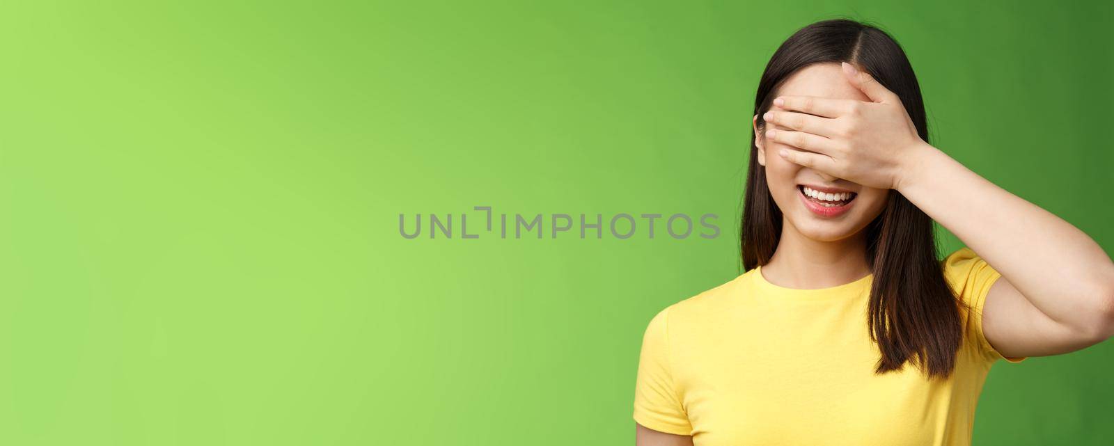 Close-up cheerful amused tender asian girlfriend close eyes waiting friend give surprise gift, smiling broadly hold palm face, anticipating interesting present, stand green background intrigued.