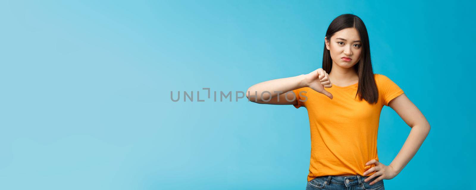 Moody unsatisfied cute asian girl picky, disliking bad restaurant food smirking displeased, grimacing disappointed show thumb down, shaking head negative judgement, stand blue background.