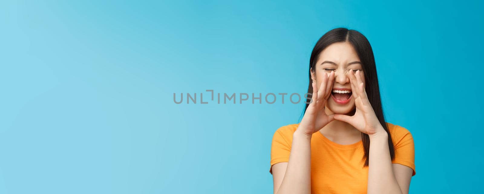 Carefree asian enthusiastic girl shouting long distance, calling friend, breaking free emotions, yelling out loud, hold hands near opene mouth rupor, searching someone in crowd, blue background.
