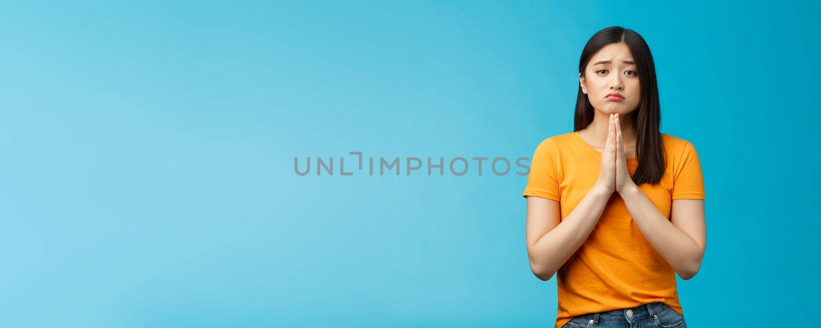 Upset cute silly asian girl praying, plead for help, pouting frowning need, make pitty face, hold hands pray begging for favor, apologizing feelings guilty sad, stand blue background.