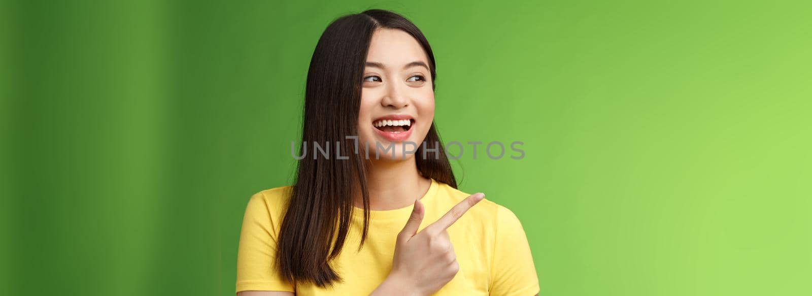 Close-up cheerful friendly asian outgoing girl having fun, pointing turn left amused, lively talking, discuss friends interesting event, stand green background chatting carefree entertained.