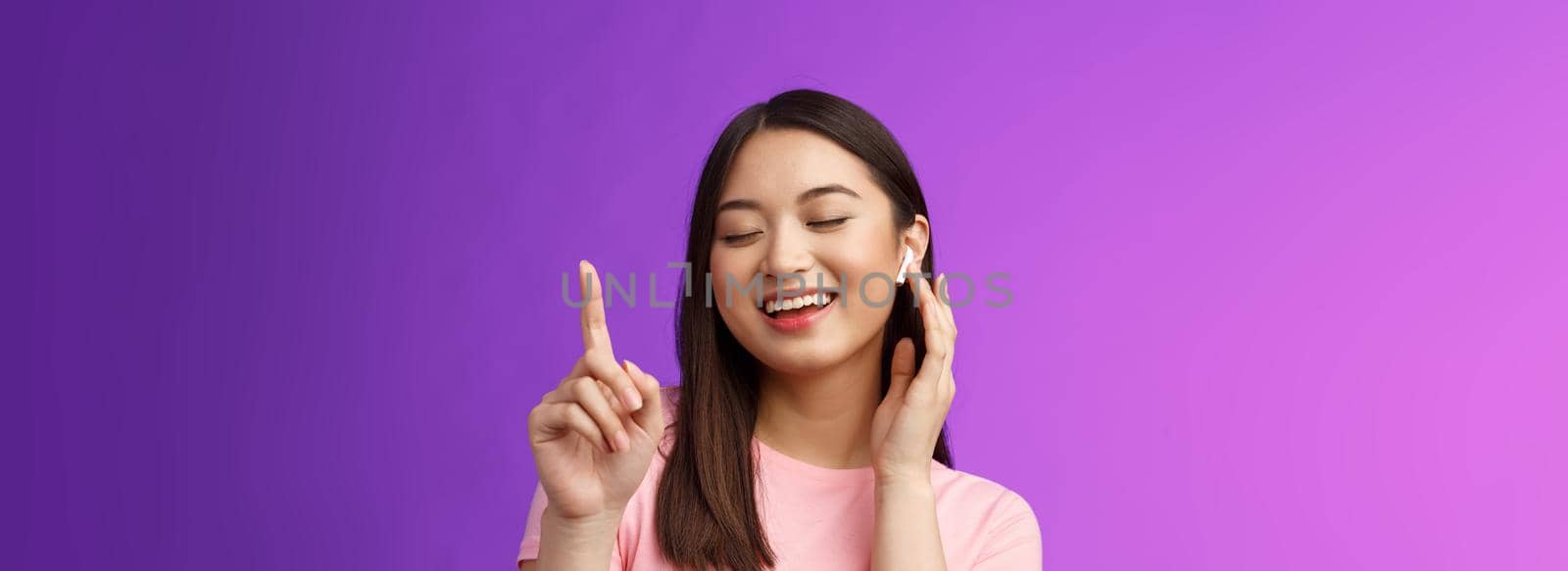 Close-up cheerful glad attractive asian woman close eyes raising one finger waiting chorus, smiling delighted touch wireless earphone, make volume louder satisfied enjoy songs, purple background.