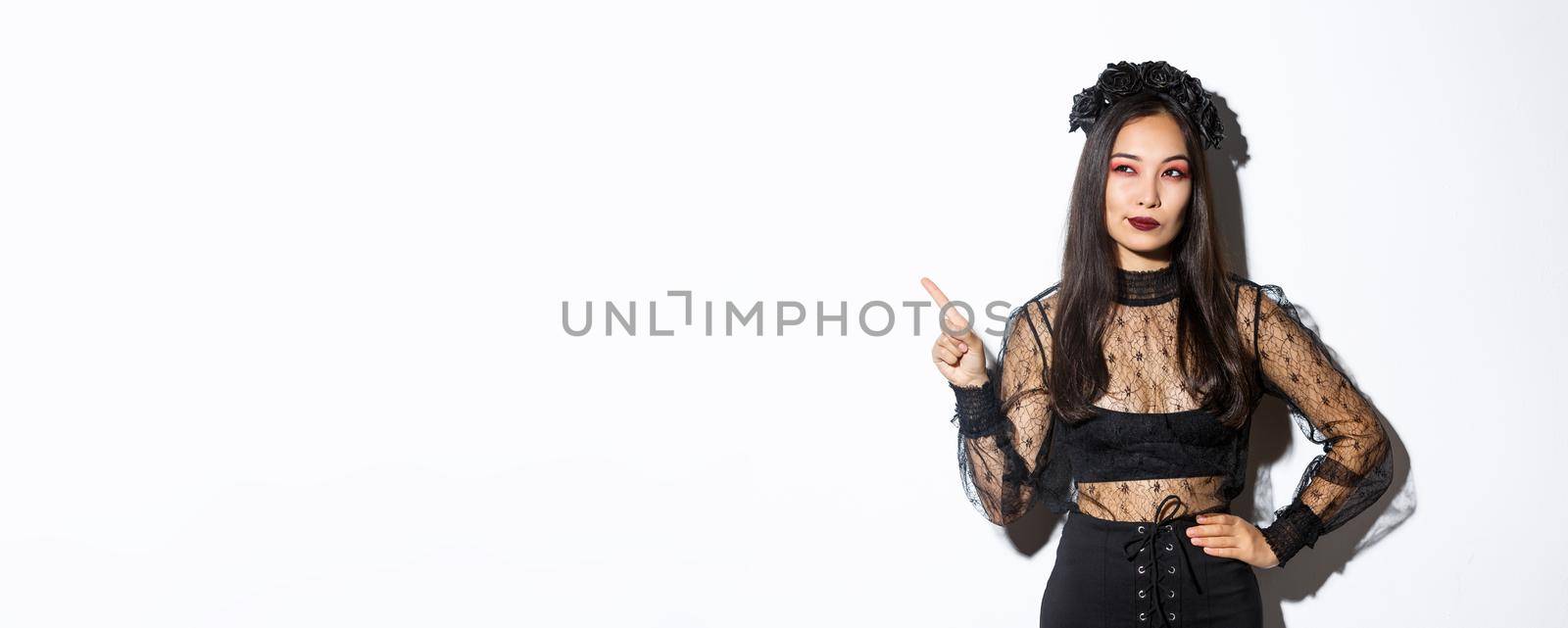 Curious attractive asian woman in witch costume, pointing finger upper left corner, looking interested in halloween promo, standing over white background and thinking.