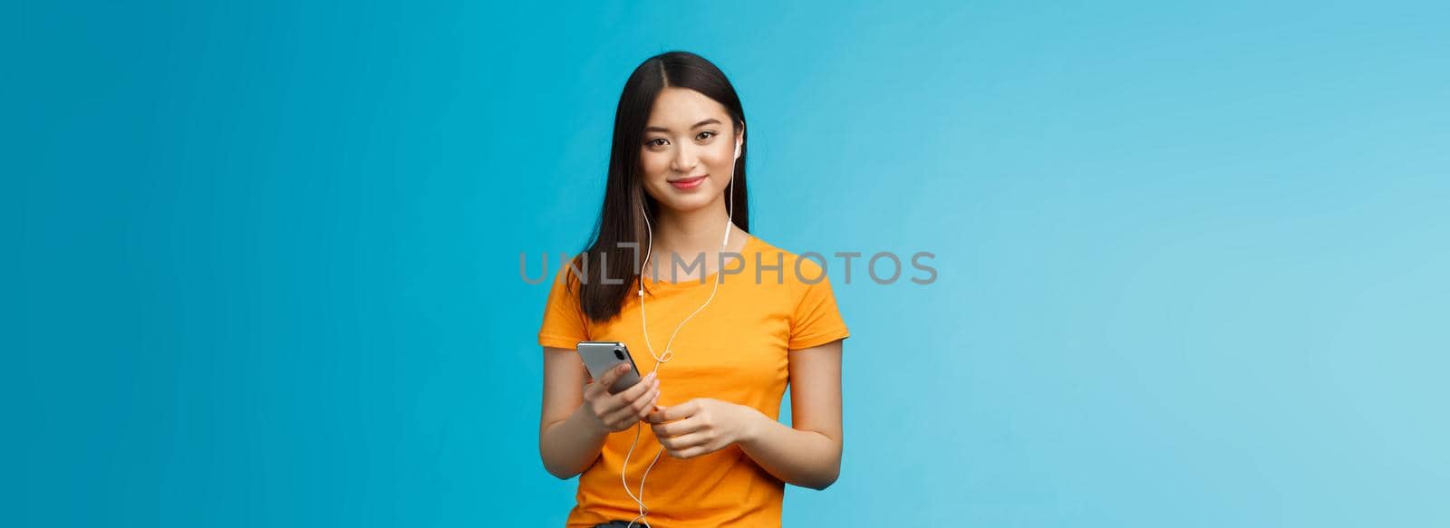 Carefree attractive pleasant, tender asian urban woman wear earphones, listen music via headphones hold smartphone, smiling delighted, found awesome new song, stand blue background.
