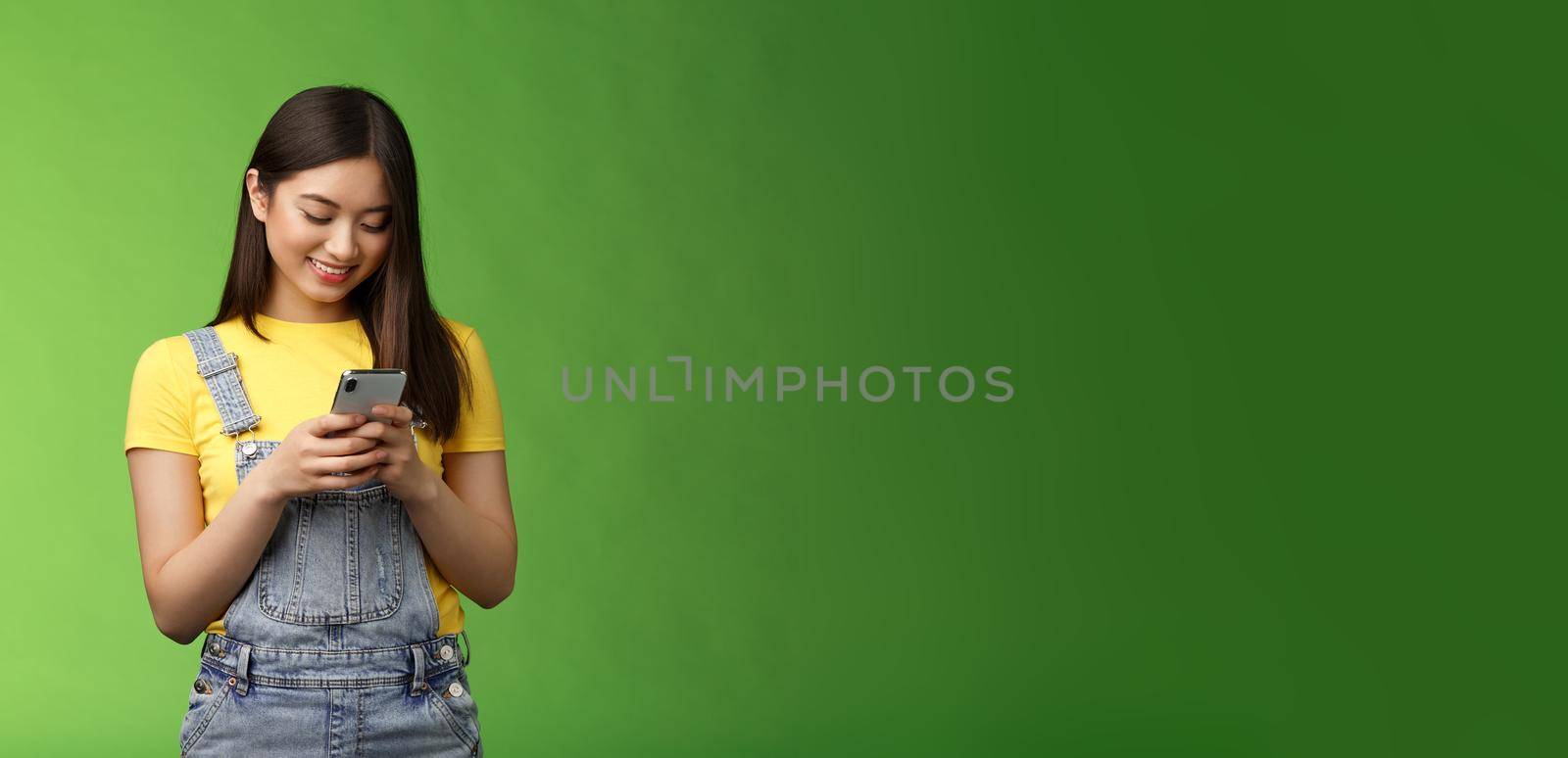 Outgoing cute asian brunette woman scroll online shop, purchase summer tour internet, hold smartphone, look amused telephone screen, use app, edit photos post social media, stand green background by Benzoix