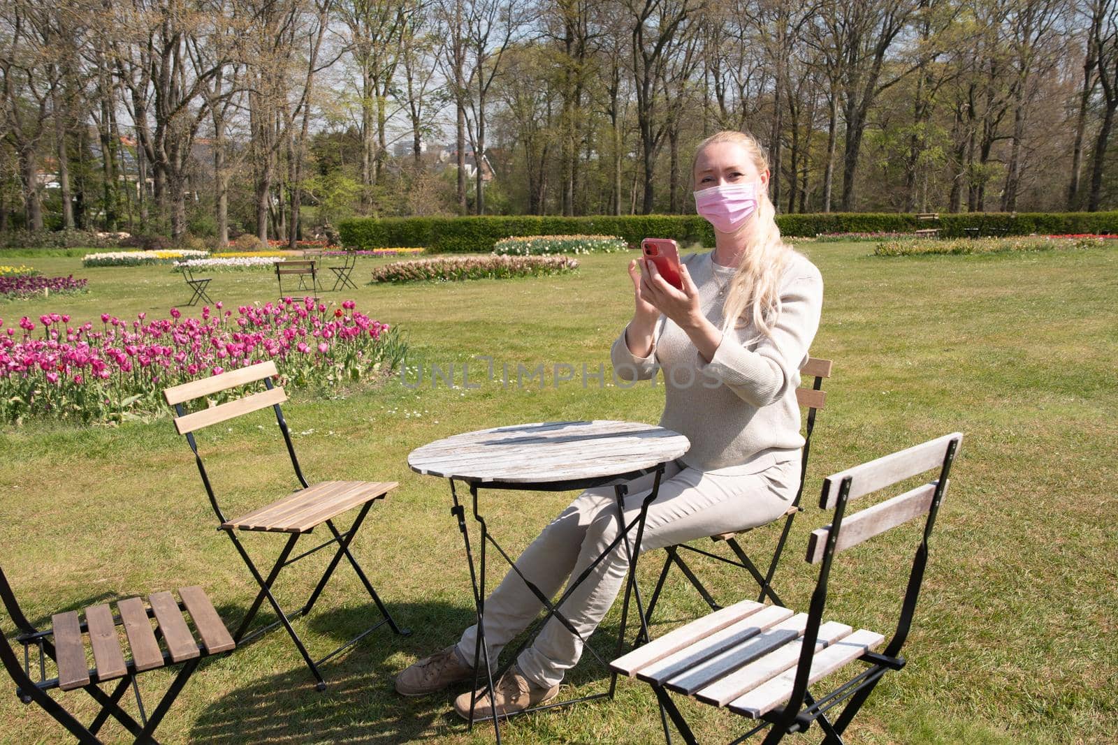 young woman in a beige jumper and a protective mask takes a selfie in the park against the backdrop of blooming tulips in early spring social media using a smartphone, High quality photo