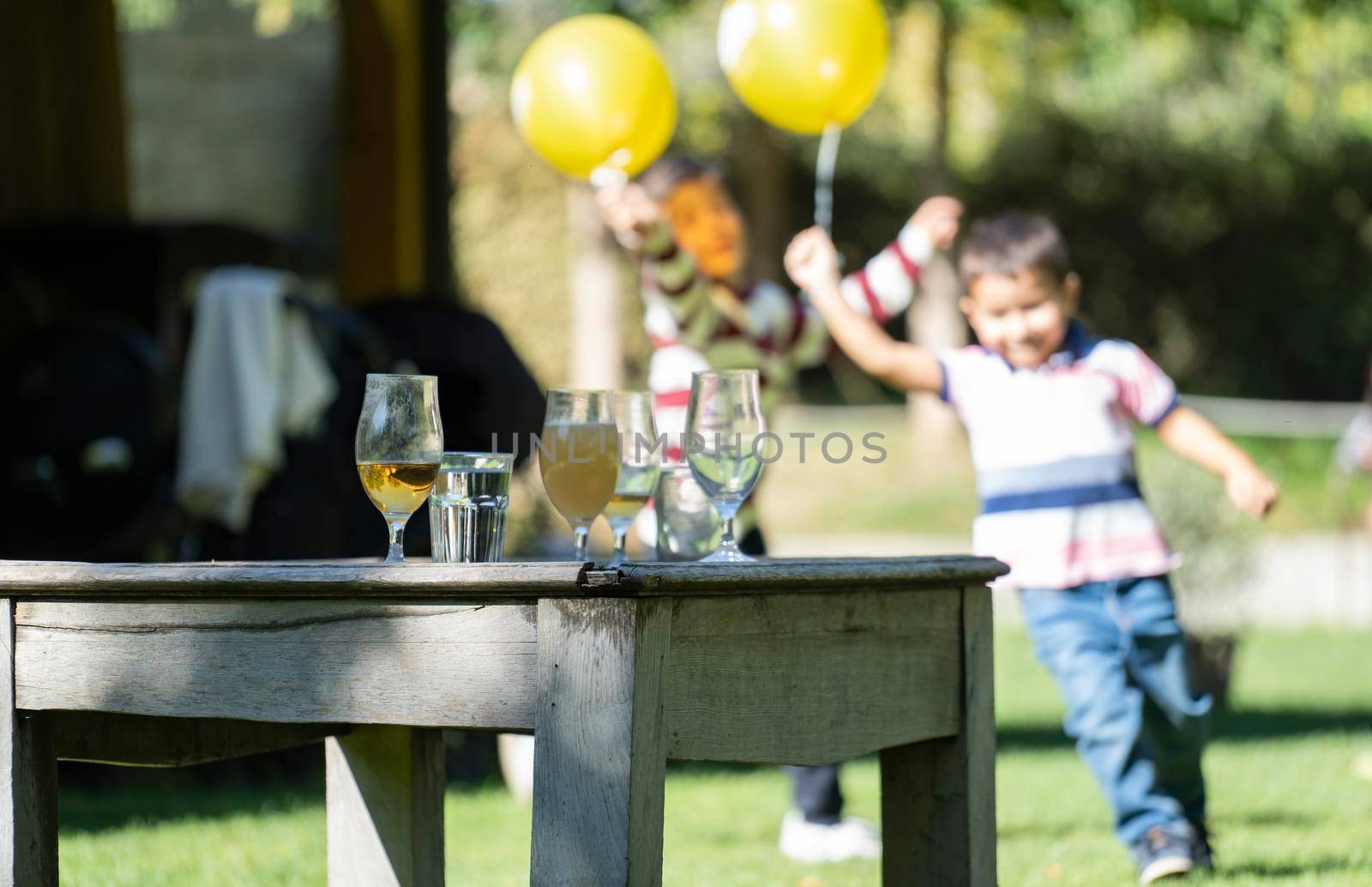 Glasses with beer and water on a vintage table during a party. Happy children playfully running with yellow balloons on blurred background.