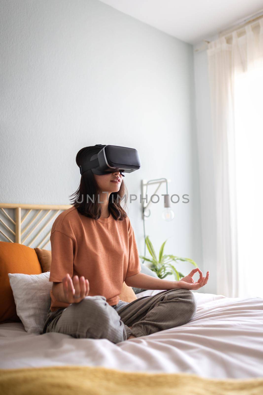 Teen asian girl meditating with help of VR experience app sitting on bed. Chinese young woman doing meditation using virtual reality goggles at home. Vertical. Copy space. Technology and healthy lifestyle concepts.