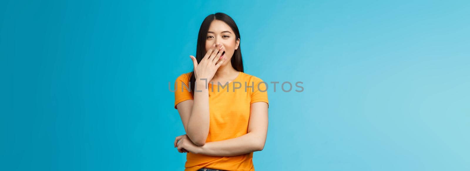 Sleepy cute asian brunette yawning waiting for cup coffee morning standing office smiling upbeat, cover opened mouth waking up feel energized, stand blue background. Copy space