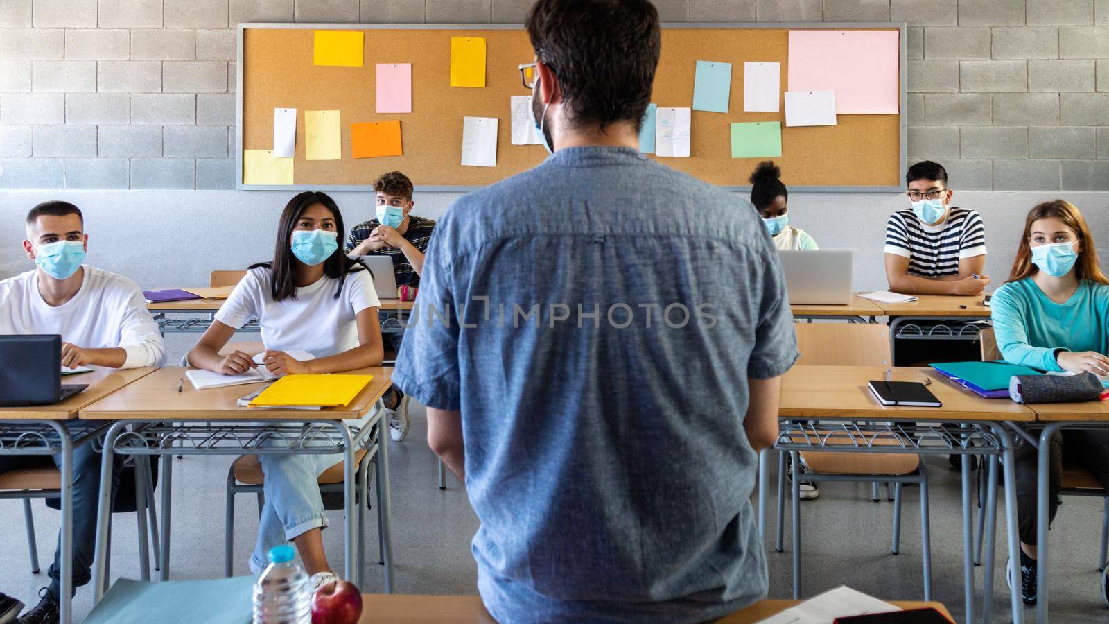 Group of multiracial teen high school students wearing face mask listen to teacher lesson. Rear view of teacher. Education concept. Healthcare concept.