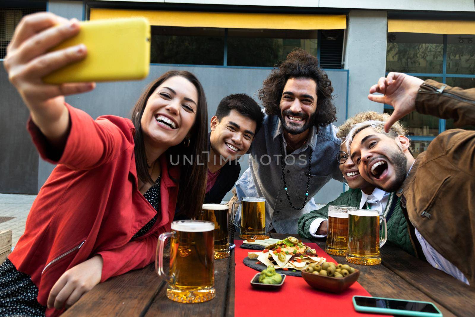 Happy multiracial group of friends enjoying beer and food together take selfie with cell phone in a bar outdoors. by Hoverstock