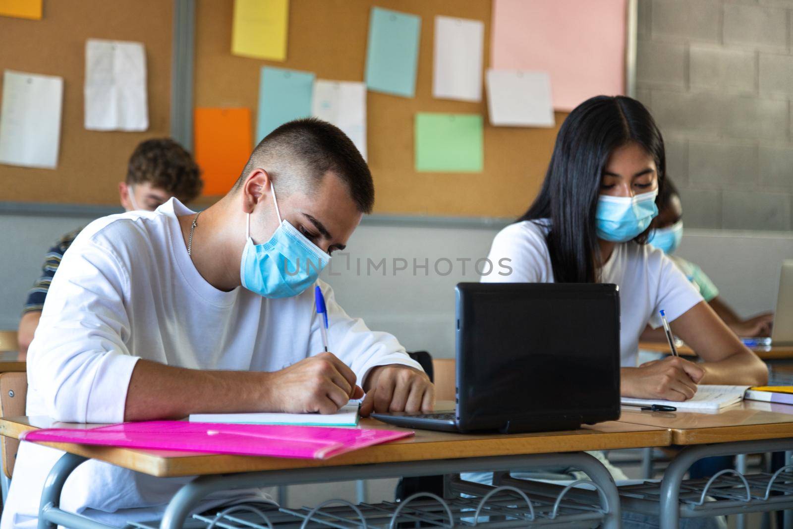 Multiracial high school students wearing a face mask in class handwriting, taking notes. by Hoverstock