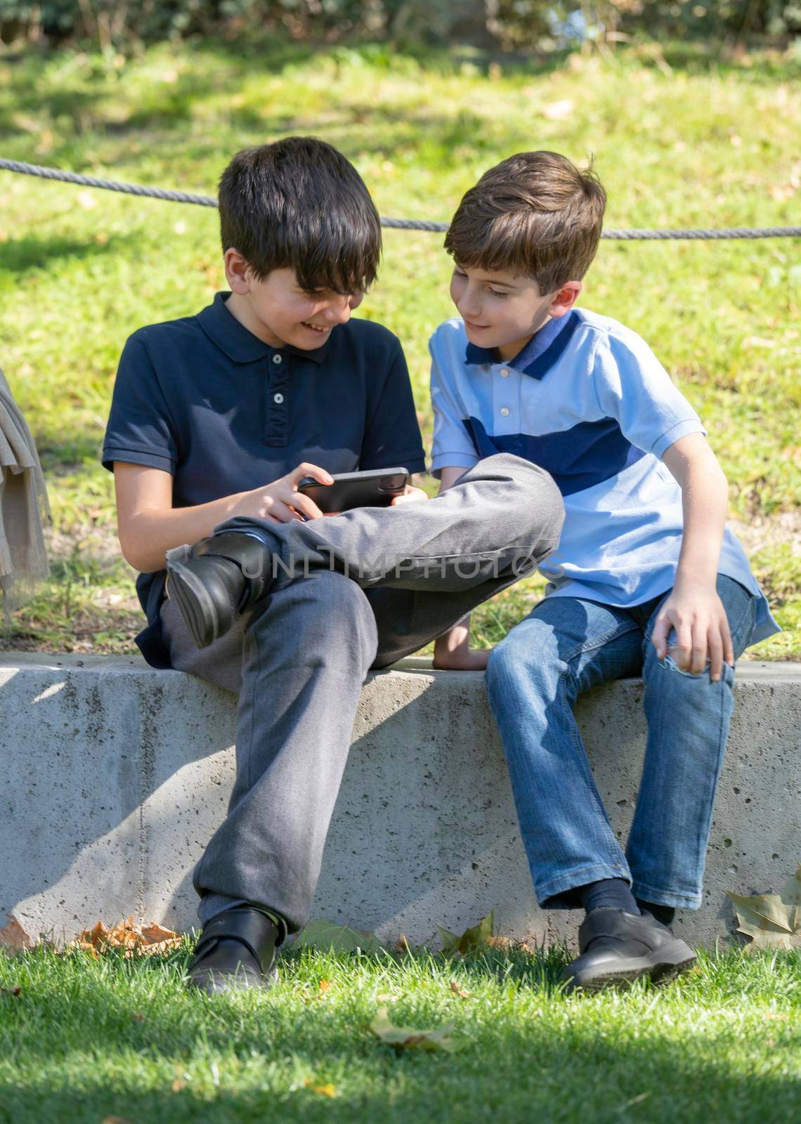 Cute two boys sitting on the park and play online games. One boy holding the smartphone. Young boys use the phone by papatonic