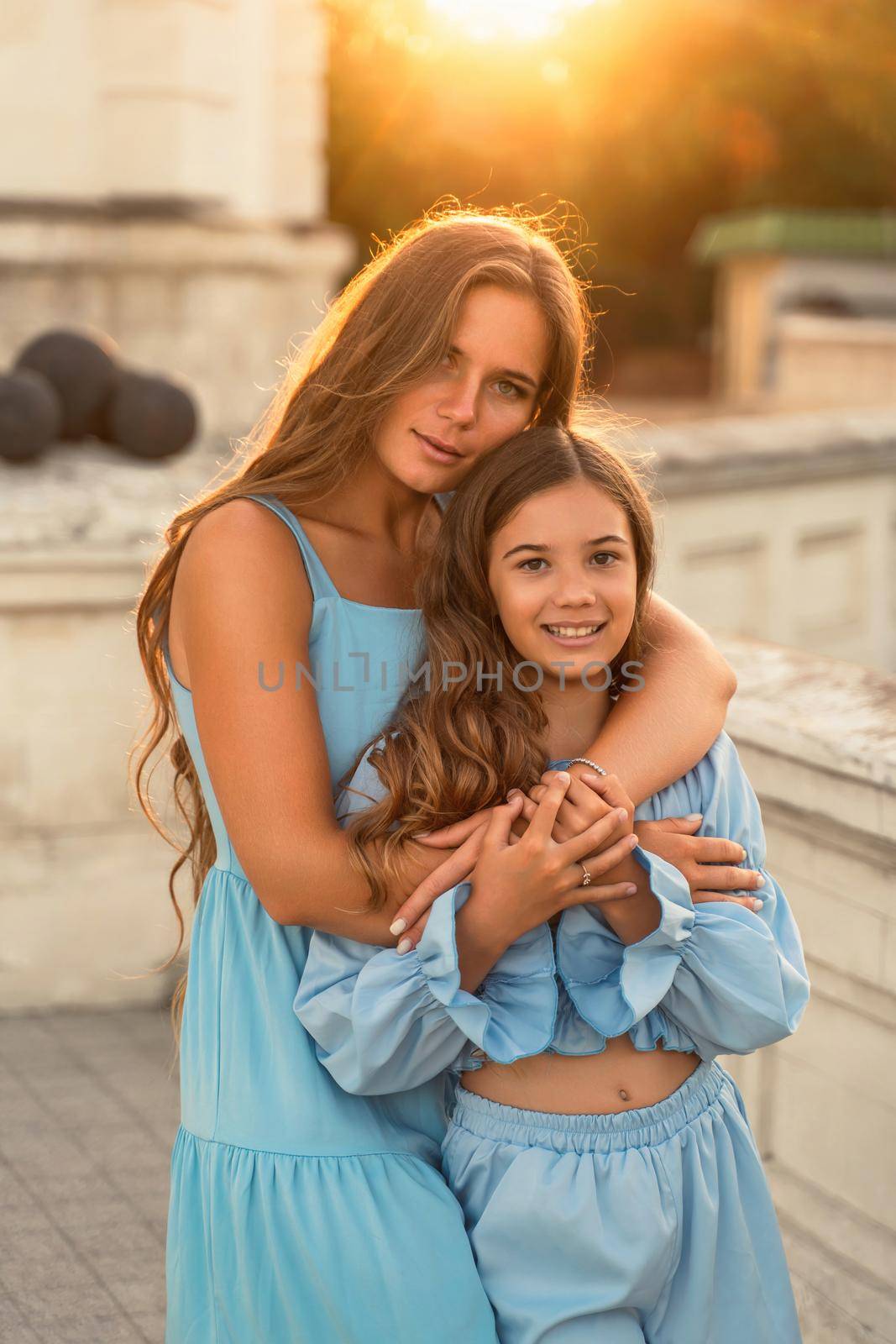 Portrait of mother and daughter in blue dresses with flowing long hair against the backdrop of sunset. The woman hugs and presses the girl to her. They are looking at the camera