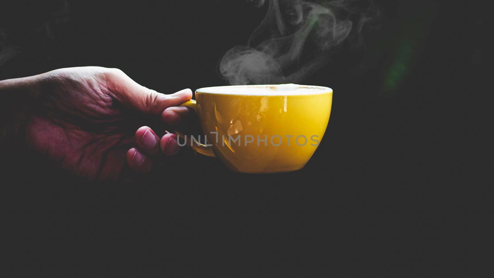 Close-up isolated hand holding yellow mug on black background. Hand hold simple yellow cup. Perfect for both tea and coffee.