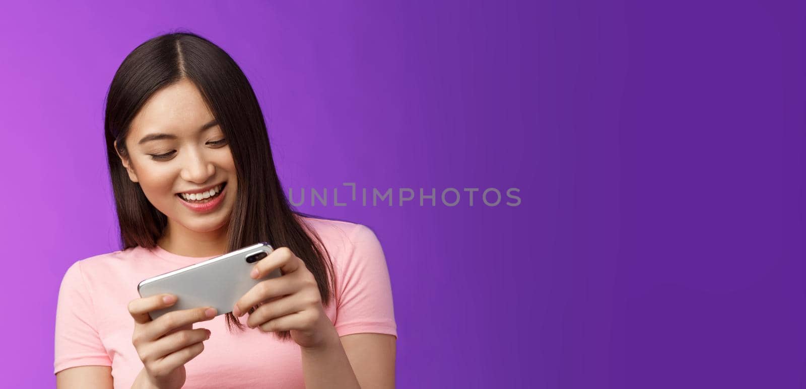 Close-up joyful attractive asian woman brunette having fun spend time playing smartphone game, laughing smiling eager win race, hold phone horizontal beating score, purple background.