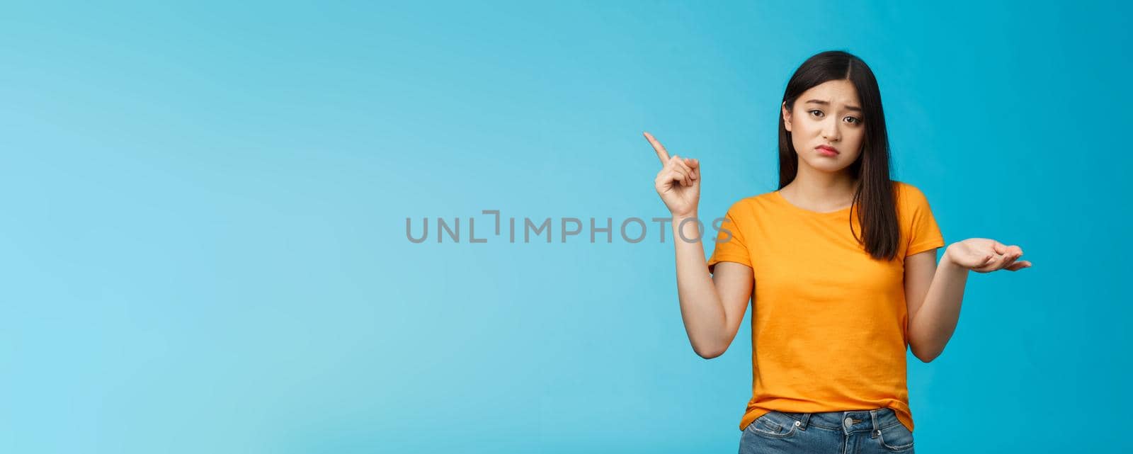 Troubled cute upset asian girl have problem asking your help, pointing upper left corner shrugging unsure, look frustrated and sad, questioned answer, cannot deal problem, stand blue background.