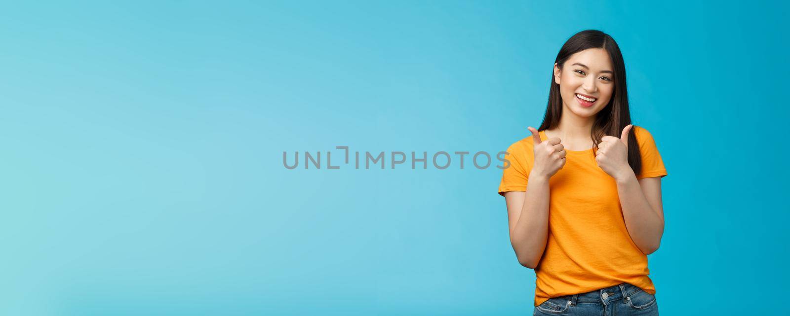 Supportive cute friendly asian girl give thumbs up and smiling, cheering for you, congratulate nice work, approving good idea, like nice outfit, grinning glad accept plan, stand blue background.