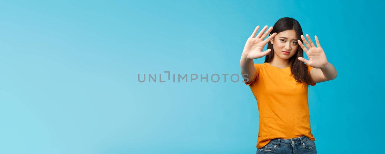 Asian moody displeased silly timid girl asking turn off light not photograph her, raise hands block, cover face step back and cringe disappointed, standing blue background upset reluctant. Copy space